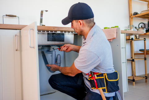 Plumber in a cap and gray shirt working under a kitchen sink, repairing pipes with a wrench, and wearing a tool belt.